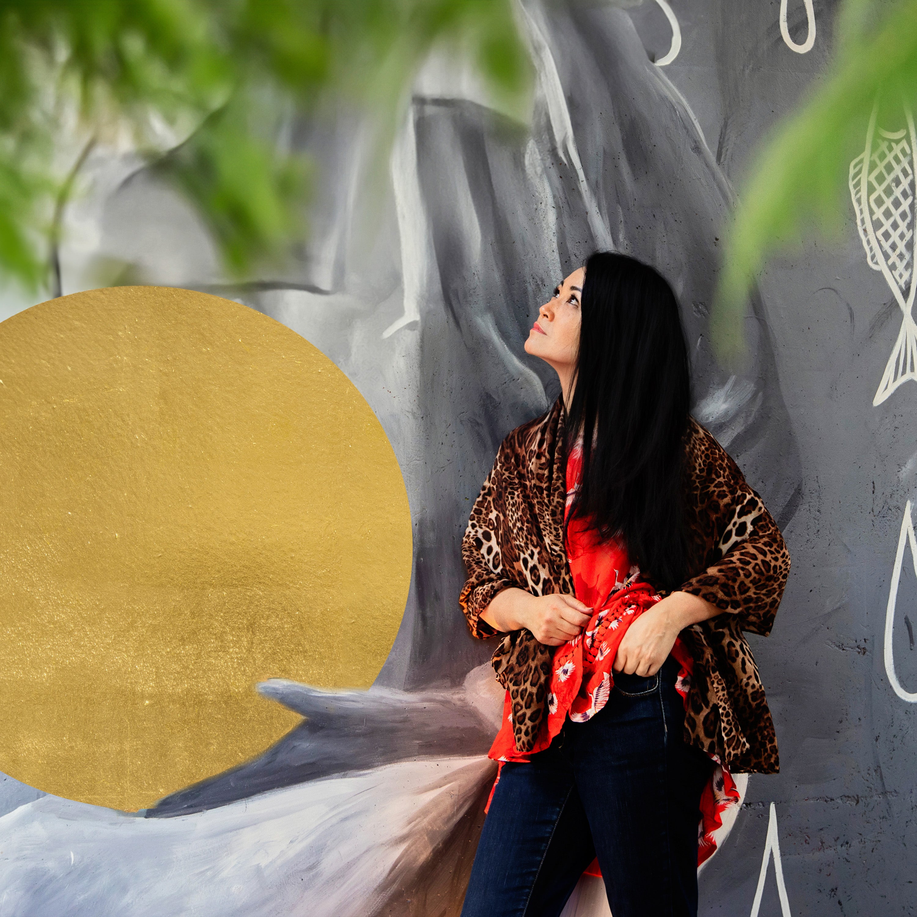 Vietnamese woman with long black hair leaning against an art mural. She wears jeans with a red top and leopard print scarf wrap.