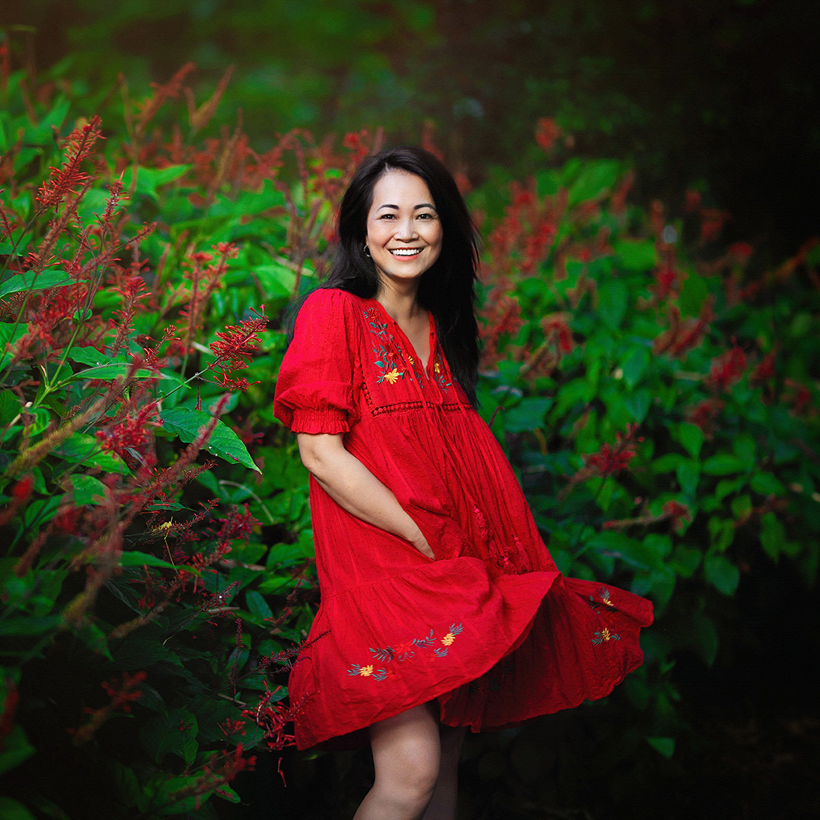 Smiling Vietnamese woman with hands in the pockets of her red dress stands near a green bush with red flowers.