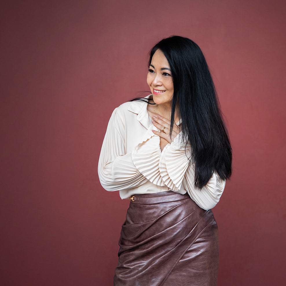  Vietnamese woman smiles and looks sideways, holding her hands to her chest. She wears a brown leather skirt and cream top against a maroon background.