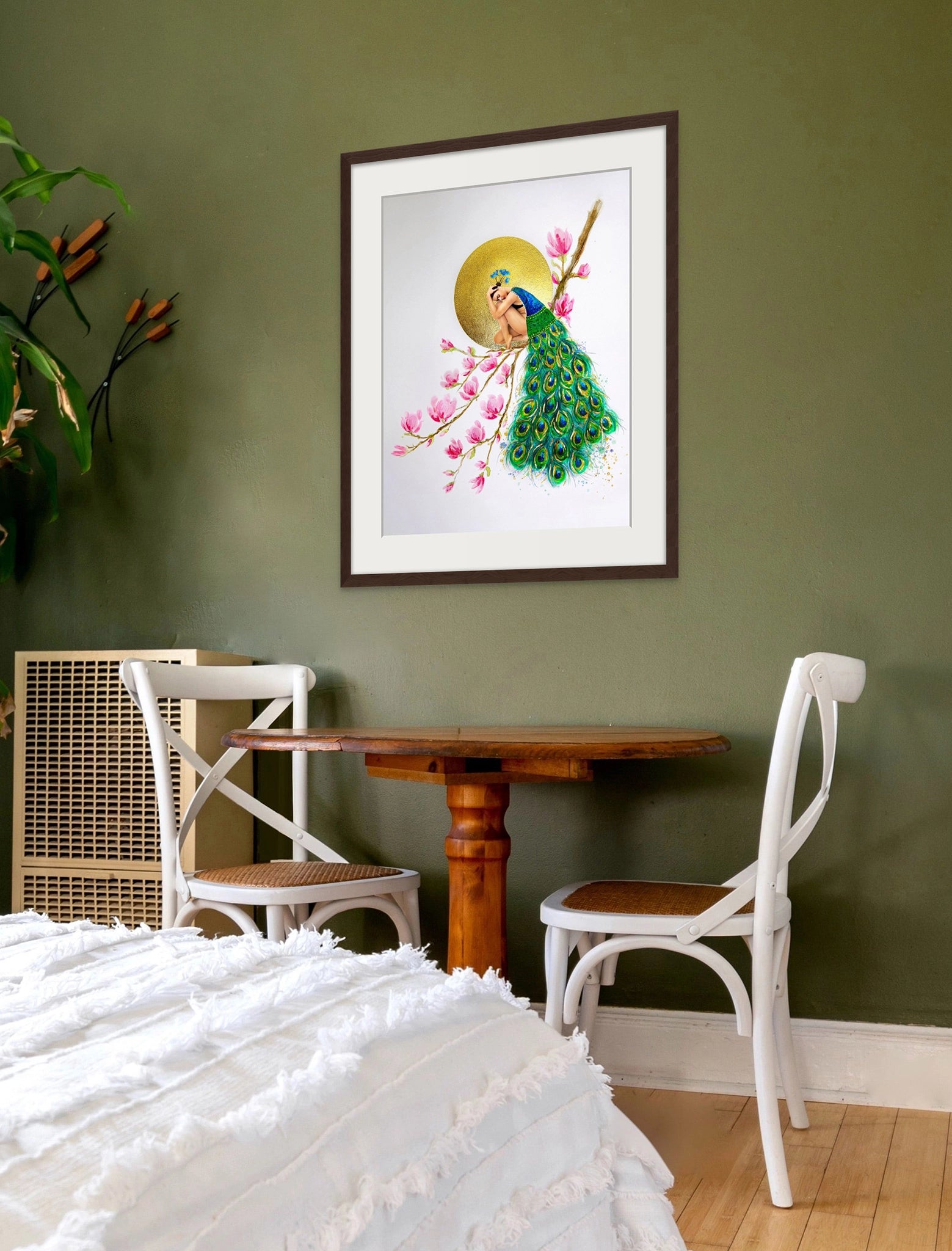 Framed Hue the Artist art print of woman in a peacock feather cloak, hangs in a bedroom with green walls, white bed spread and wood table with two chairs.