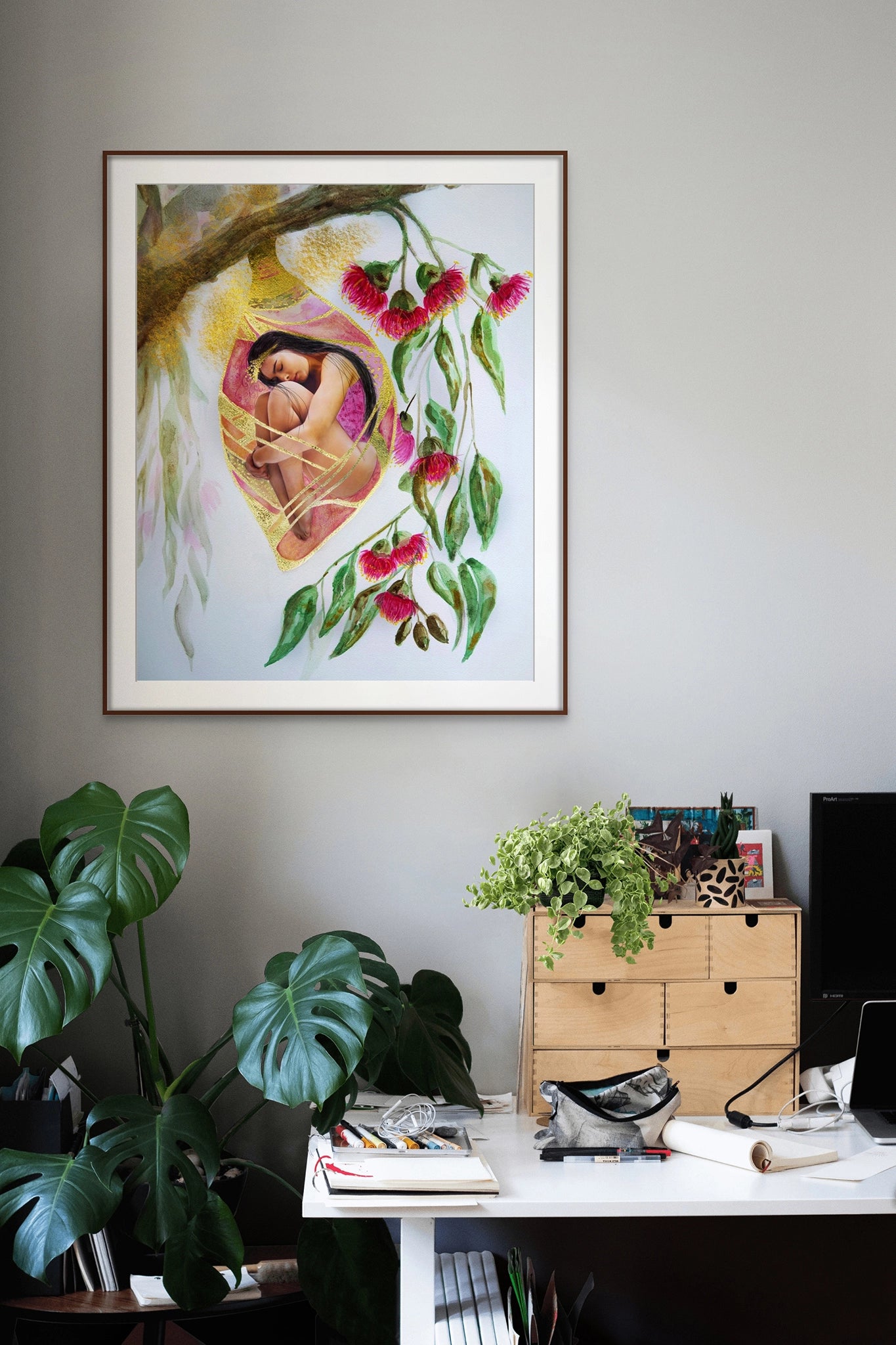 Framed Hue the Artist canvas print of woman in cocoon with pink gum nut flowers, hangs on wall of an office with monstera plant, desk, pens, pencils and computer.