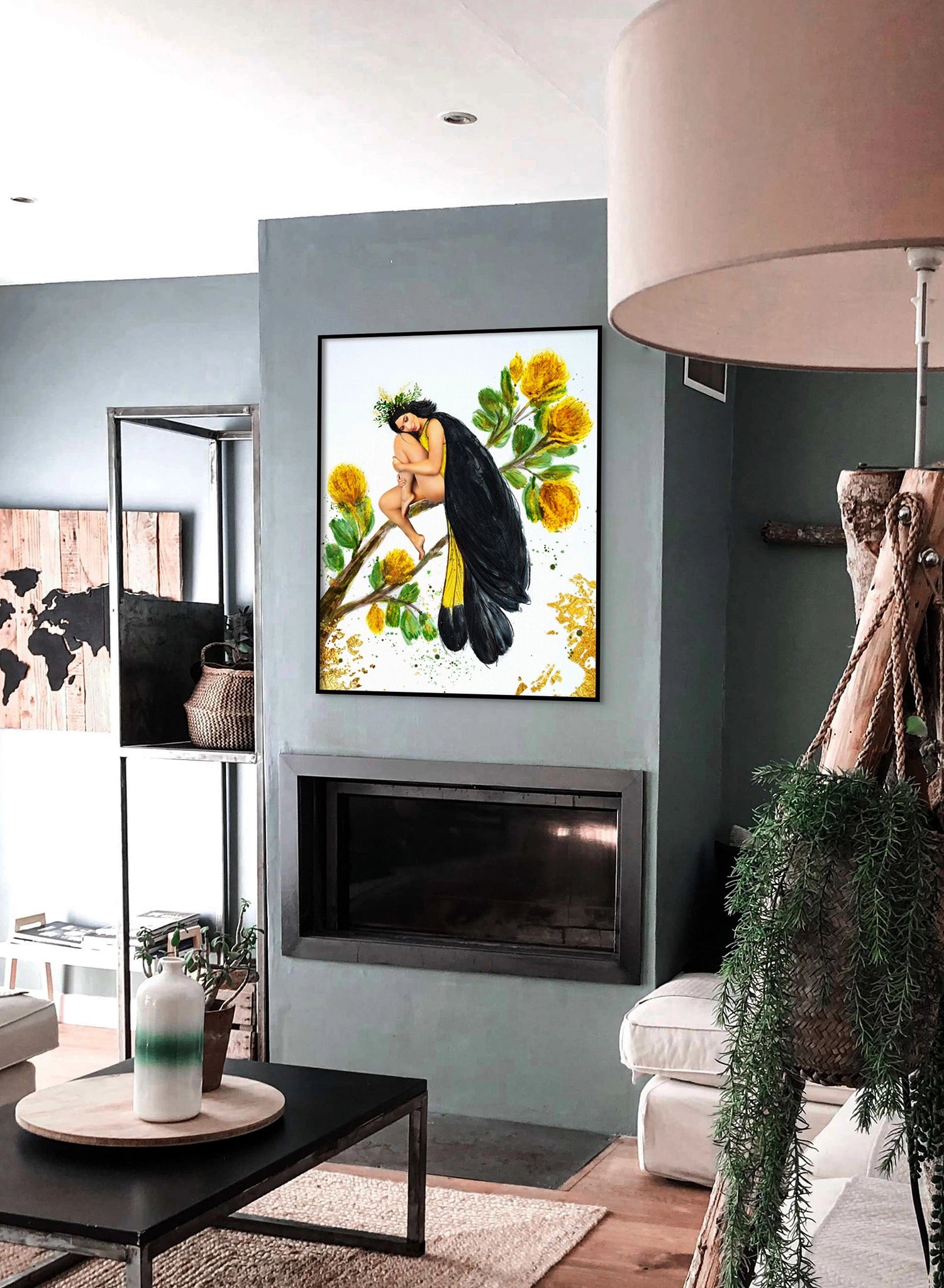 Framed Hue the Artist print of a bird-like woman in a black and yellow cloak hangs above a fireplace in a sage green living room with a lamp, coffee table, map, and potted plant.