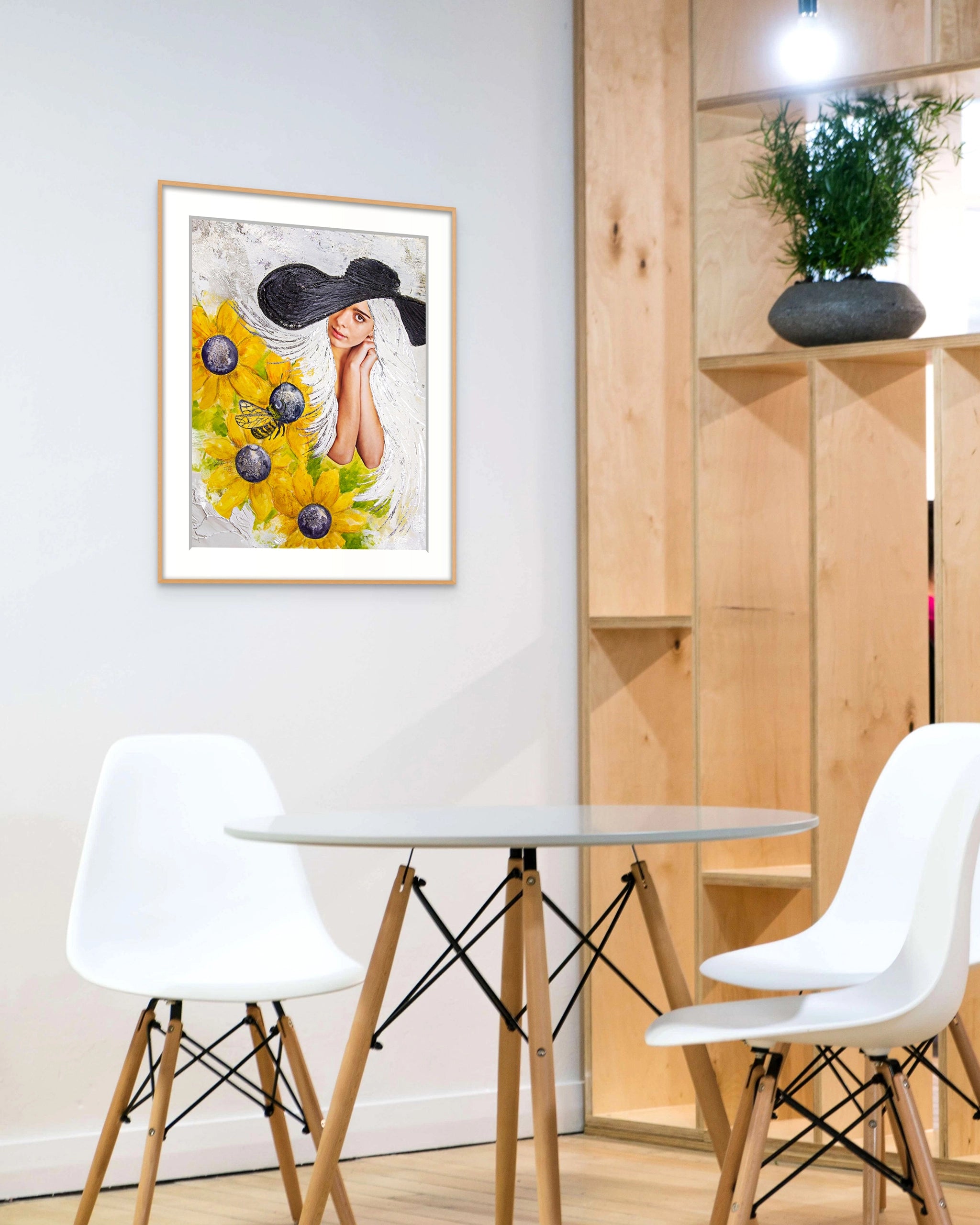 Framed Hue the Artist wall print of white haired woman wearing black floppy bat and sunflowers hangs above a small table with three white chairs. 