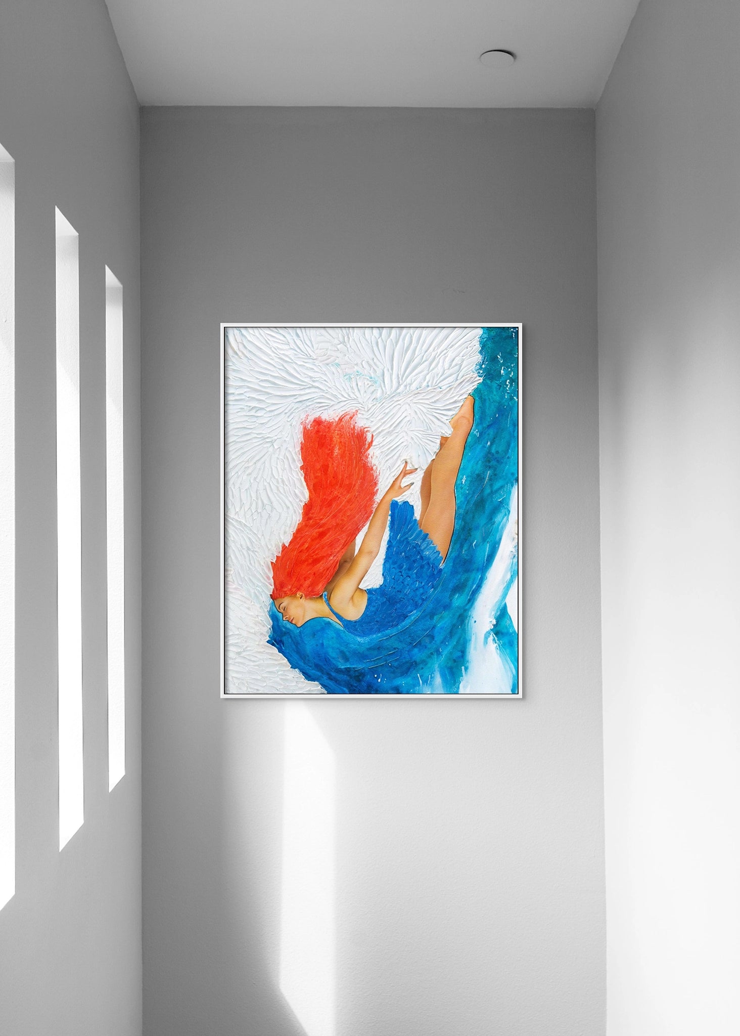 Framed artwork by Hue the Artist of a red-haired woman diving into a blue and white ocean hangs in a tall stairwell with light streaming through three narrow windows.