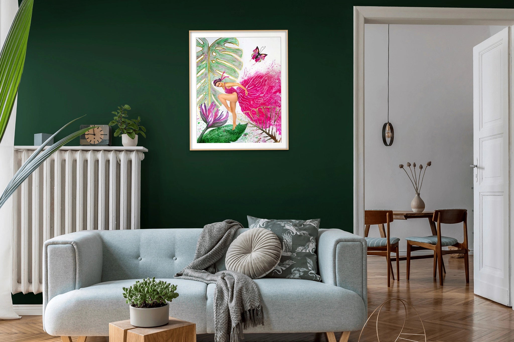 Framed Hue the Artist art print of woman in a bright pink dress hangs on a green wall. Under it is a light blue couch and a dining table with two chairs is visible in the next room.