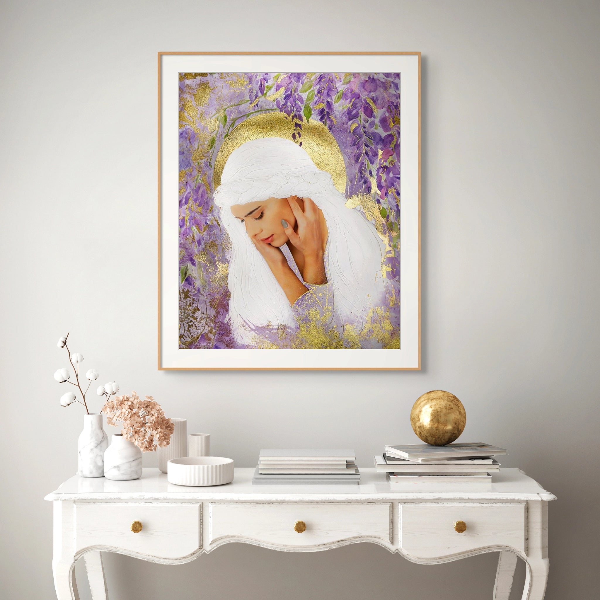 Framed Hue the Artist art print of a sad white haired woman, surrounded by purple wisteria and gold foil details hangs above a white hall desk with books and decorative white pots.